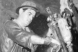 A black and white image shows a man working underground in a mine. He is wet with sweat.