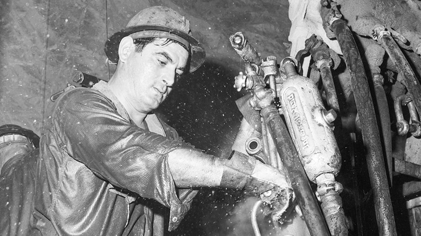 A black and white image shows a man working underground in a mine. He is wet with sweat.