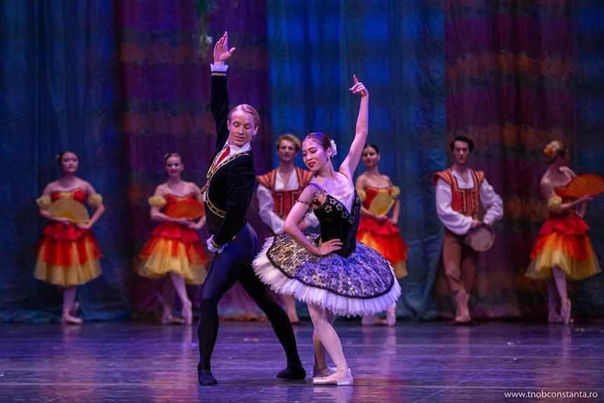 A male ballet dancer next to a ballerina posed on stage in front of a chorus.