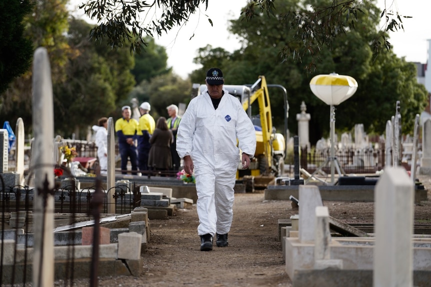 A man in white overalls walks past gravestones as excavation crews work in the background