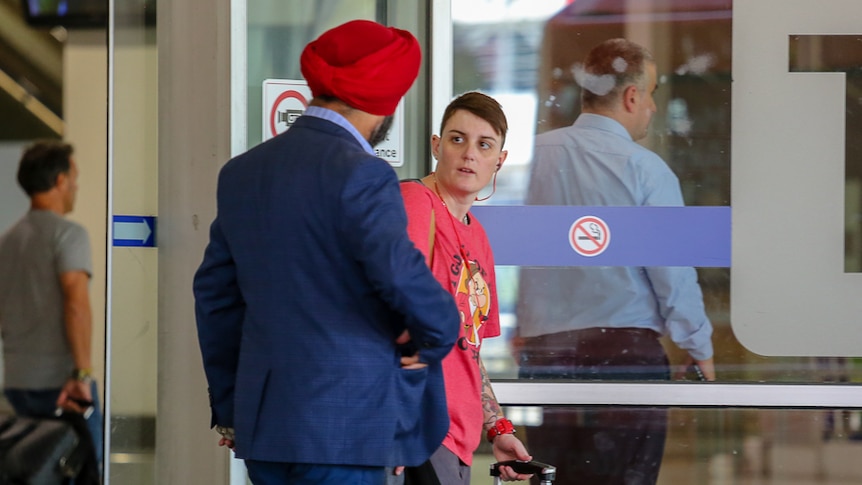 A taxi driver in a suit approaches a person with a red shirt and a suitcase outside an airport.