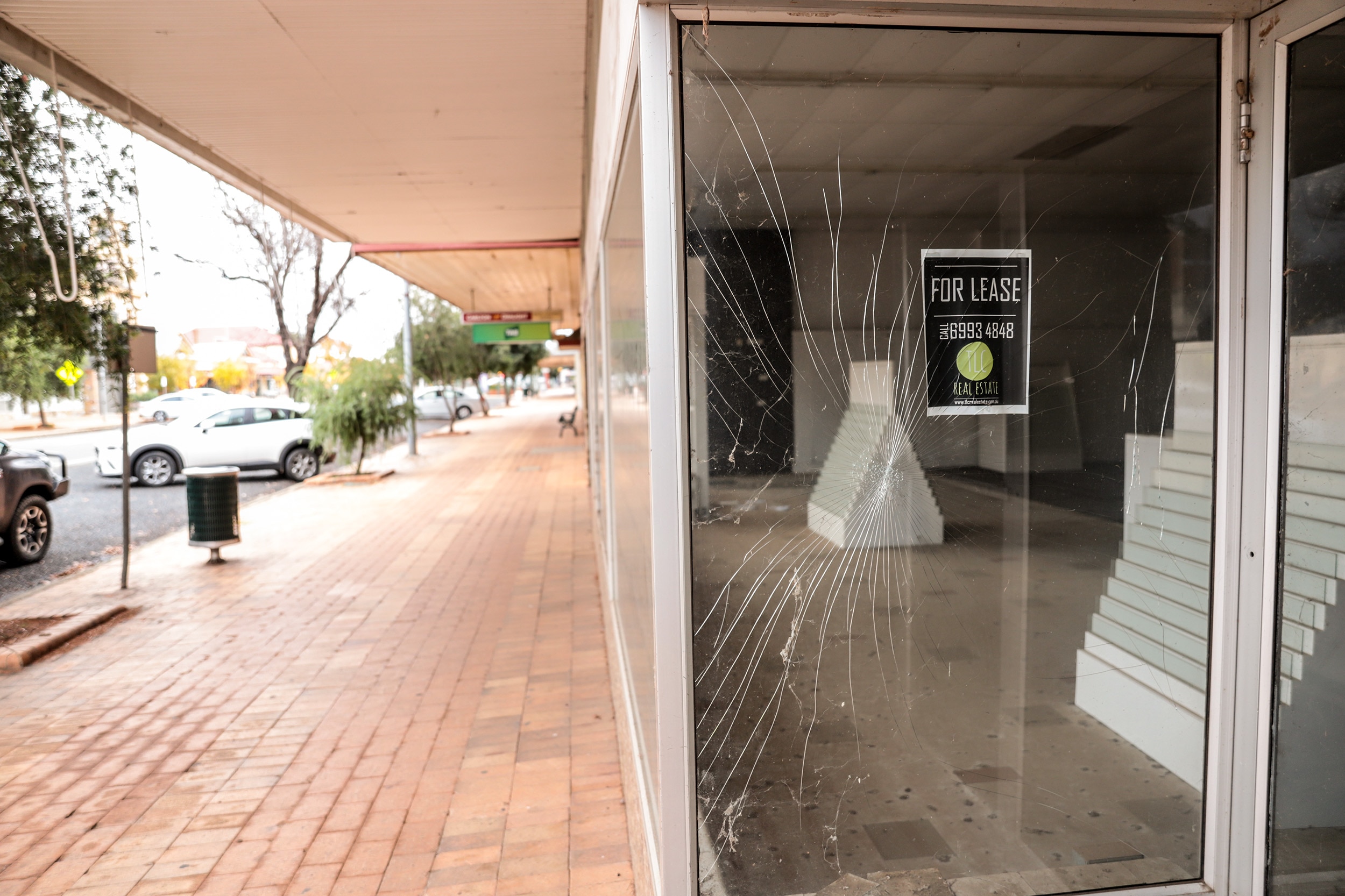 Shattered glass in the window of a shop next to a sign reading 'For Lease'. There is a footpath and parked cars to the left.