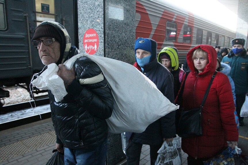 Worried-looking people wearing warm clothing and carrying sacks of their belongings walk along a train platform.