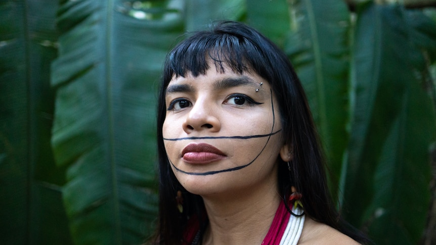A close up of Txai Suruí, a woman with black hair and a blunt fringe. She has thick black line traditional tattoos on her face.