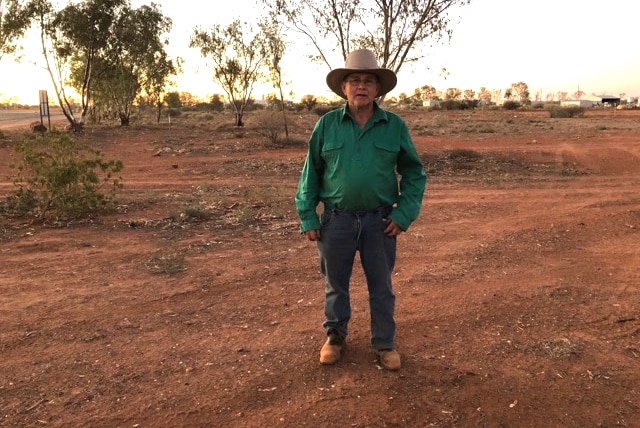 Mayor of Bulloo Shire John Ferguson stands in paddock with red dirt wearing green shirt, jeans and wide brimmed hat