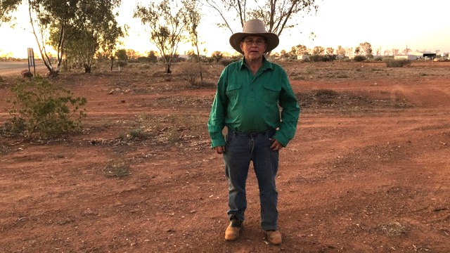 Mayor of Bulloo Shire John Ferguson stands in paddock with red dirt wearing green shirt, jeans and wide brimmed hat