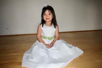 Young girl wearing tiny tiara and white dress sits on floor smiling with hands neatly together.