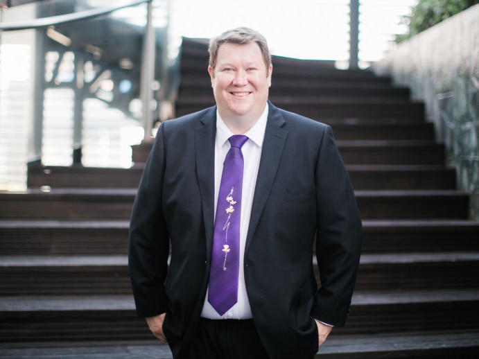 A man wearing a suit and purple tie smiles for the camera at the bottom of a staircase