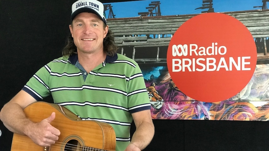 Josh Arnold stands in front of the ABC Radio Brisbane sign with his guitar. He's smiling with a thumbs up.