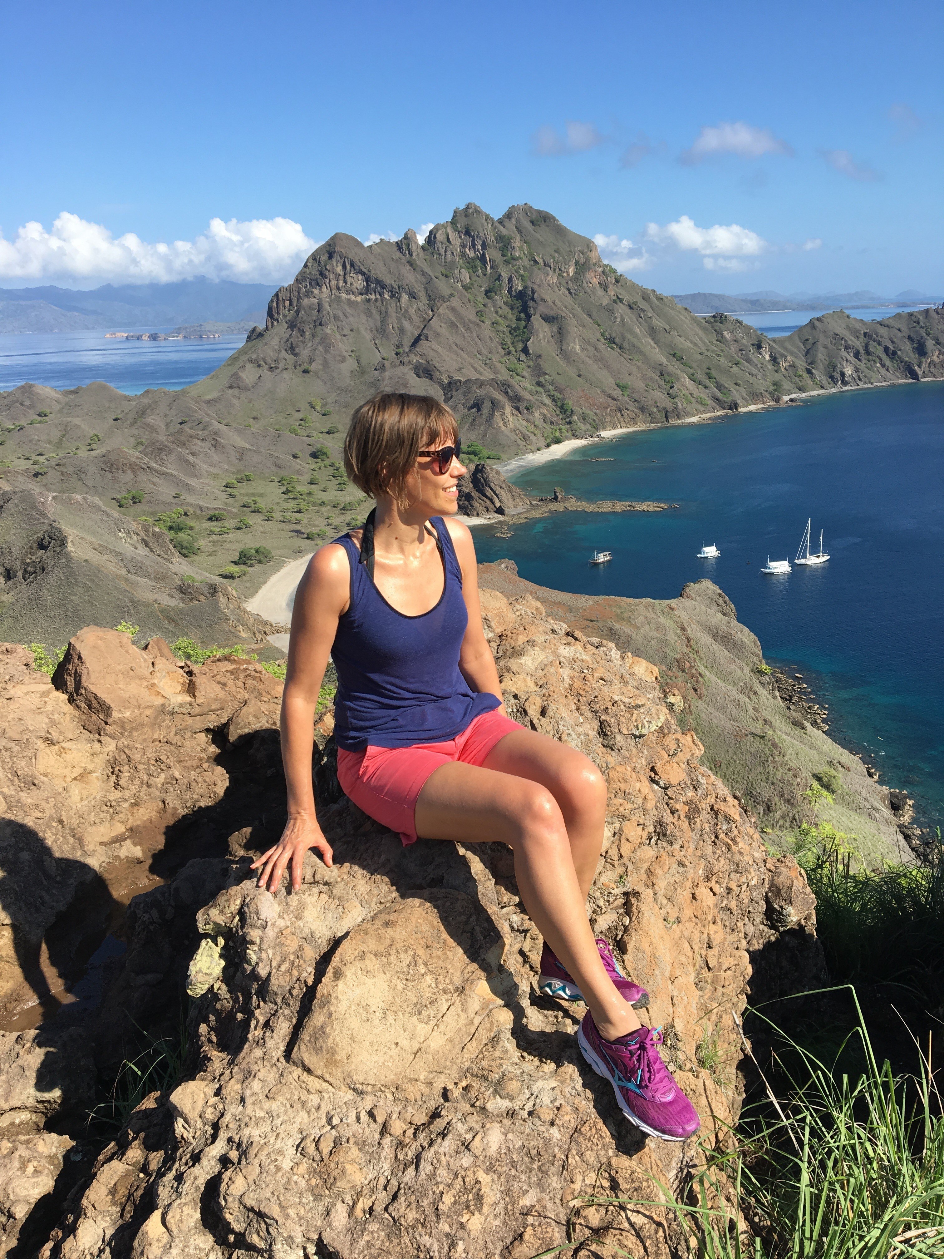 A woman wearing pink shorts, a blue singlet sitting with mountains behind her.