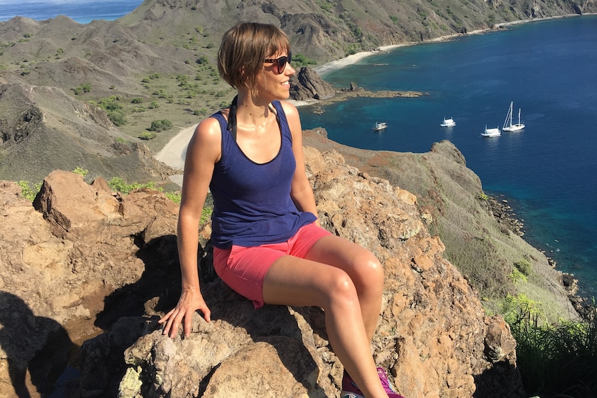 A woman wearing pink shorts, a blue singlet sitting with mountains behind her.