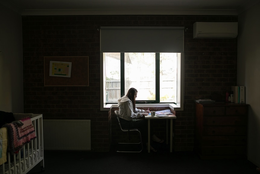 A darkened room with a woman sitting at a desk, silhouetted by a window. She is colouring-in designs on paper.