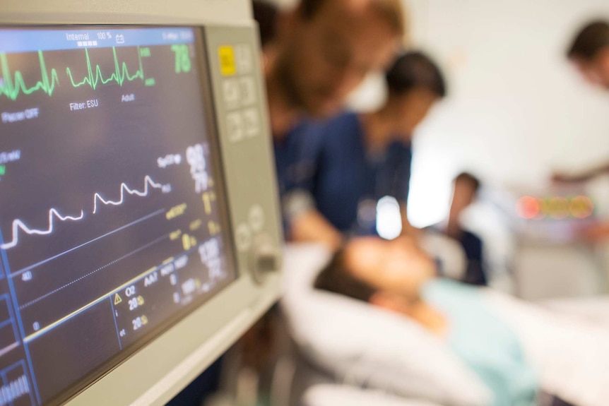 A heart rate monitor next to a person in a hospital bed with doctors around them