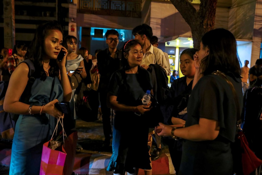 A crowd of people stand together in the street at night looking concerned.