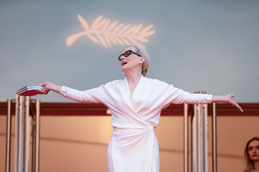 Meryl Streep in a white dress and sunglasses, arms outstretched, head thrown back, beneath the Cannes symbol