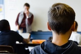 Students in a classroom with male teacher in background.