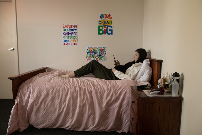 A young pregnant woman lies on a bed looking at a phone in her hand. On the wall are posters which say phrases like 'dream big'.