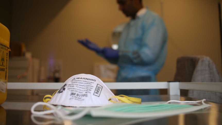 A doctor wears a blue gown and gloves in the background. A P2 mask sits in the foreground.