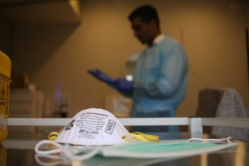 Un médecin porte une blouse bleue et des gants en arrière-plan.  Un masque P2 se trouve au premier plan.