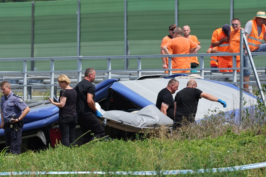 Three men in black t-shirts carry stretcher with covered grey body at crash site on hot summer's day