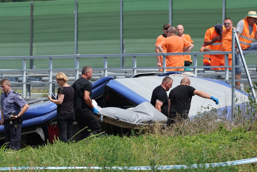Three men in black t-shirts carry stretcher with covered grey body at crash site on hot summer's day