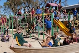 Children in maroon, green, blue and black school shirts play on a playground and old silver boat on a sunny day.