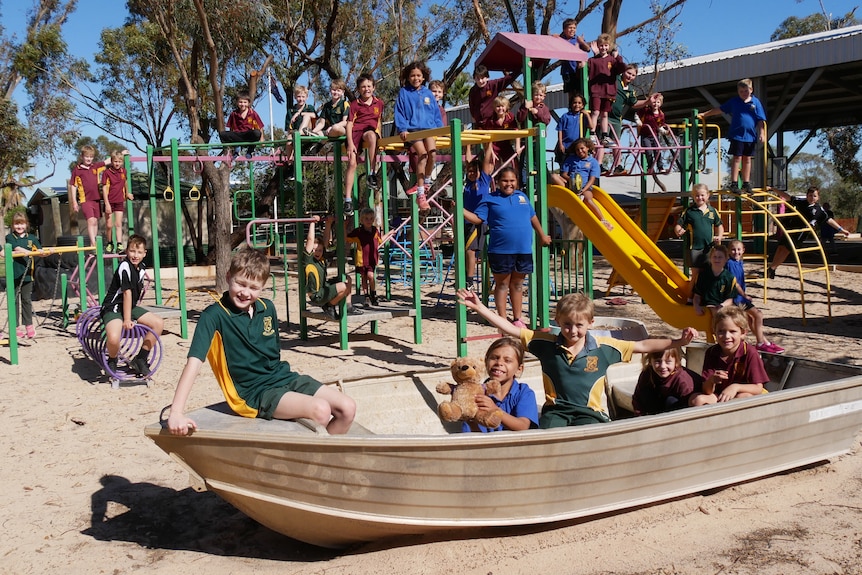 Children in maroon, green, blue and black school shirts play on a playground and old silver boat on a sunny day.