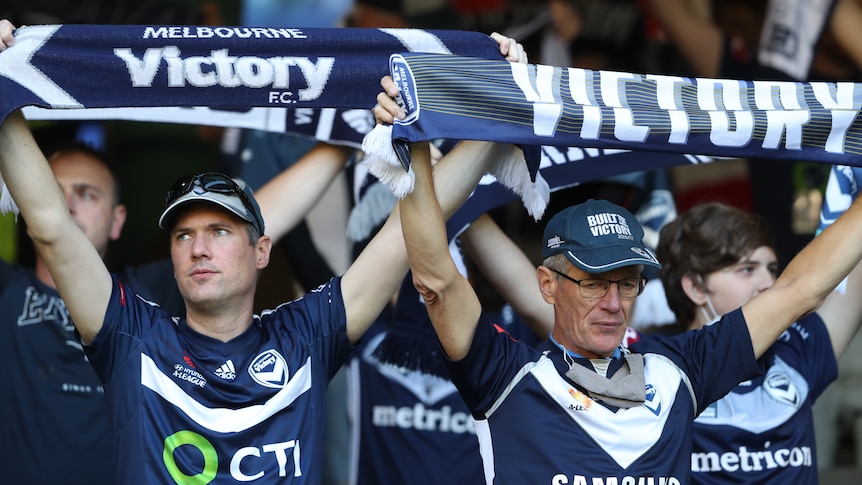 Two male Melbourne Victory fans hold up scarves saying Victory on them