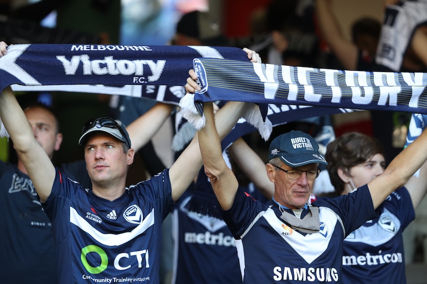 Two male Melbourne Victory fans hold up scarves saying Victory on them