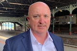 A bald man wearing a navy blue suit jacket stands on a train platform.