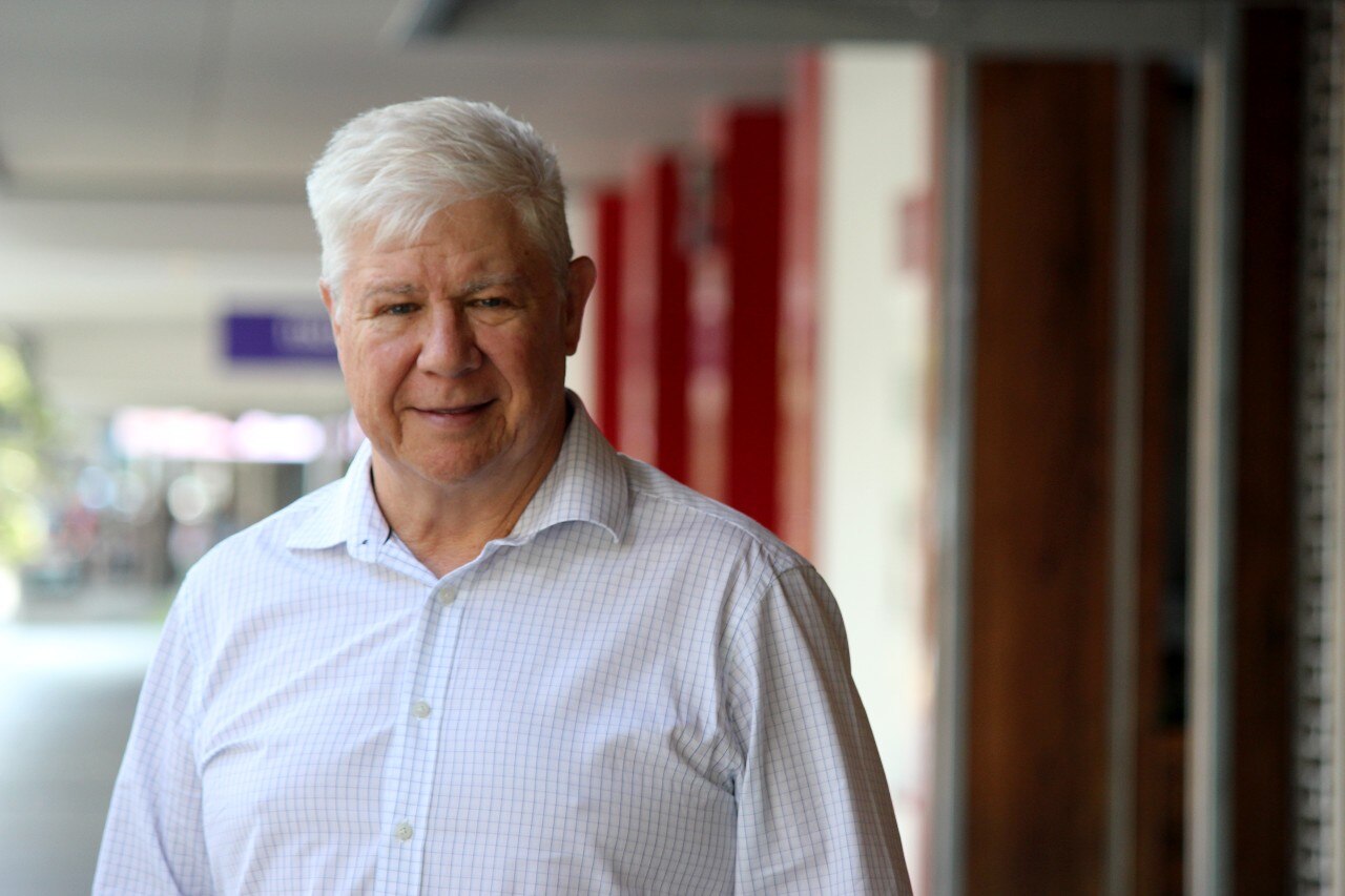 Un hombre con cabello blanco, en una calle de la ciudad, afuera. Tiene una camisa de negocios en
