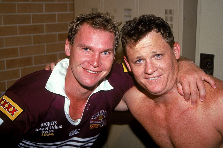 Two Queensland players - one wearing a guernsey and one without - smile at the camera after a win.