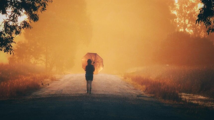 A girl walks along a morning sunlit path while holding an umbrella. The picture has an orange tinge to it
