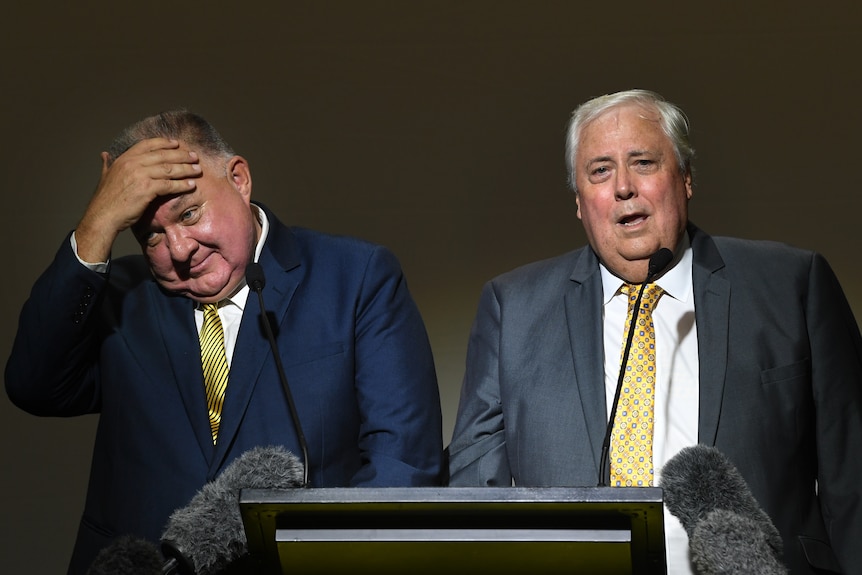 United Australia Party's Craig Kelly puts his hand to his forehead while standing on stage with Clive Palmer, who is speaking.