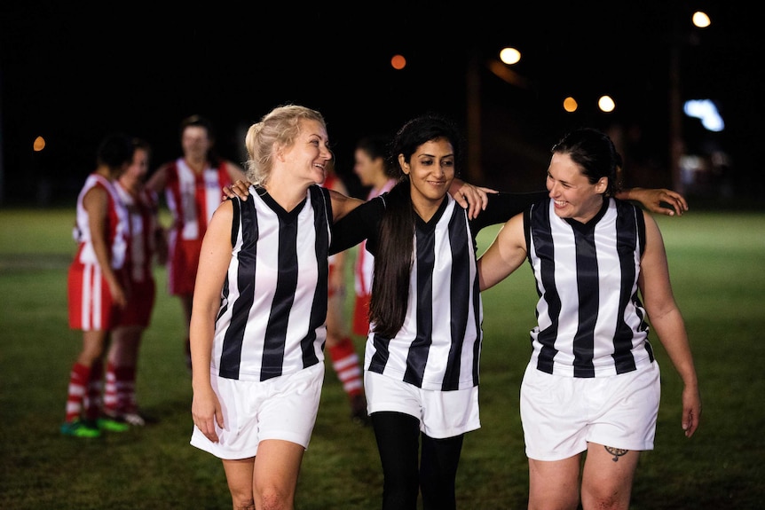 Three women wearing AFL team uniforms are walking on a football oval with their arms around each other and smiling.