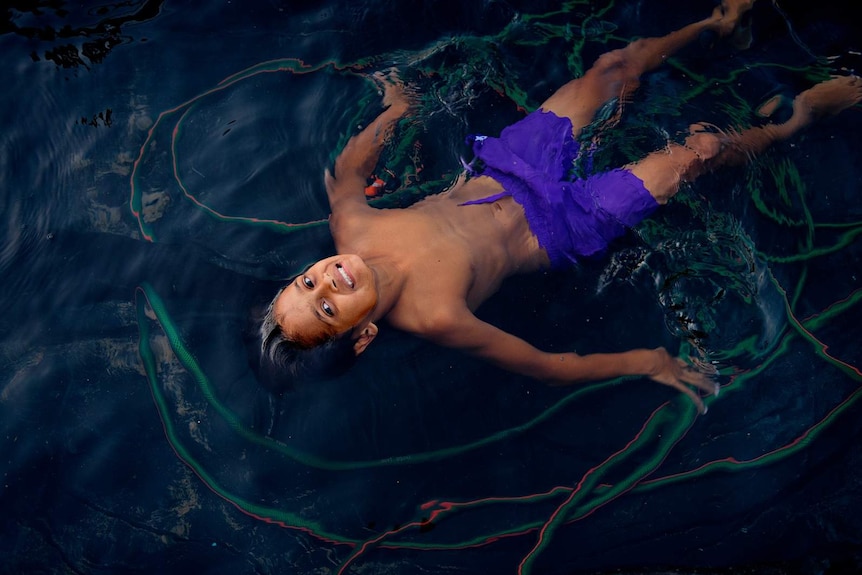 A young boy lies in an above-ground backyard pool and smiles at the camera.