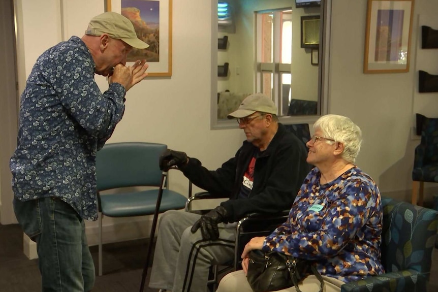 A man in a cap plays the harmonica to a man and woman who is smiling in a doctor's waiting room