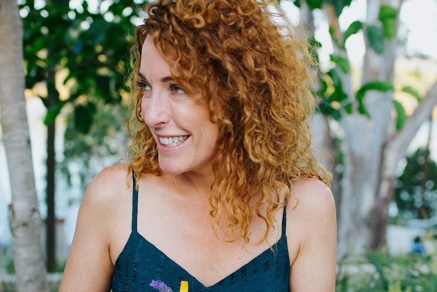 A woman with curly orange hair looking to her right smiling while holding a glass of pink gin