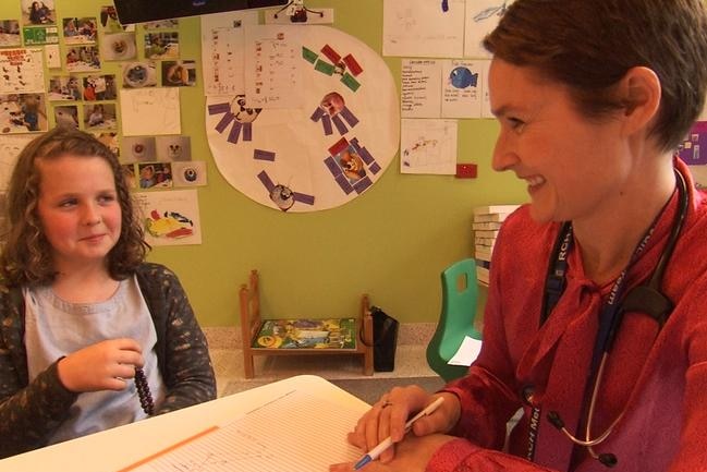 a young girl sits at a desk smiling at a female doctor who has short blonde hair