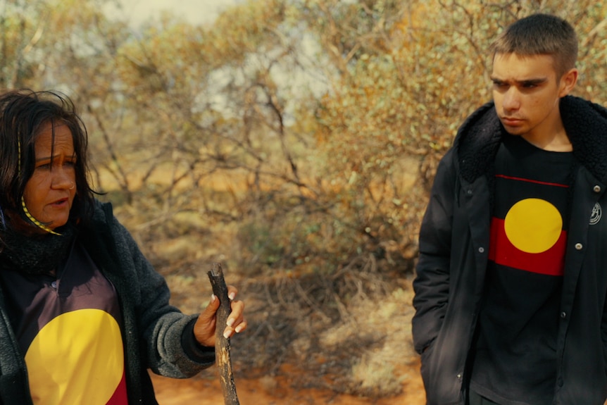 A young man looks attentively at his Nan, who is teaching him how to track emus in the bush.