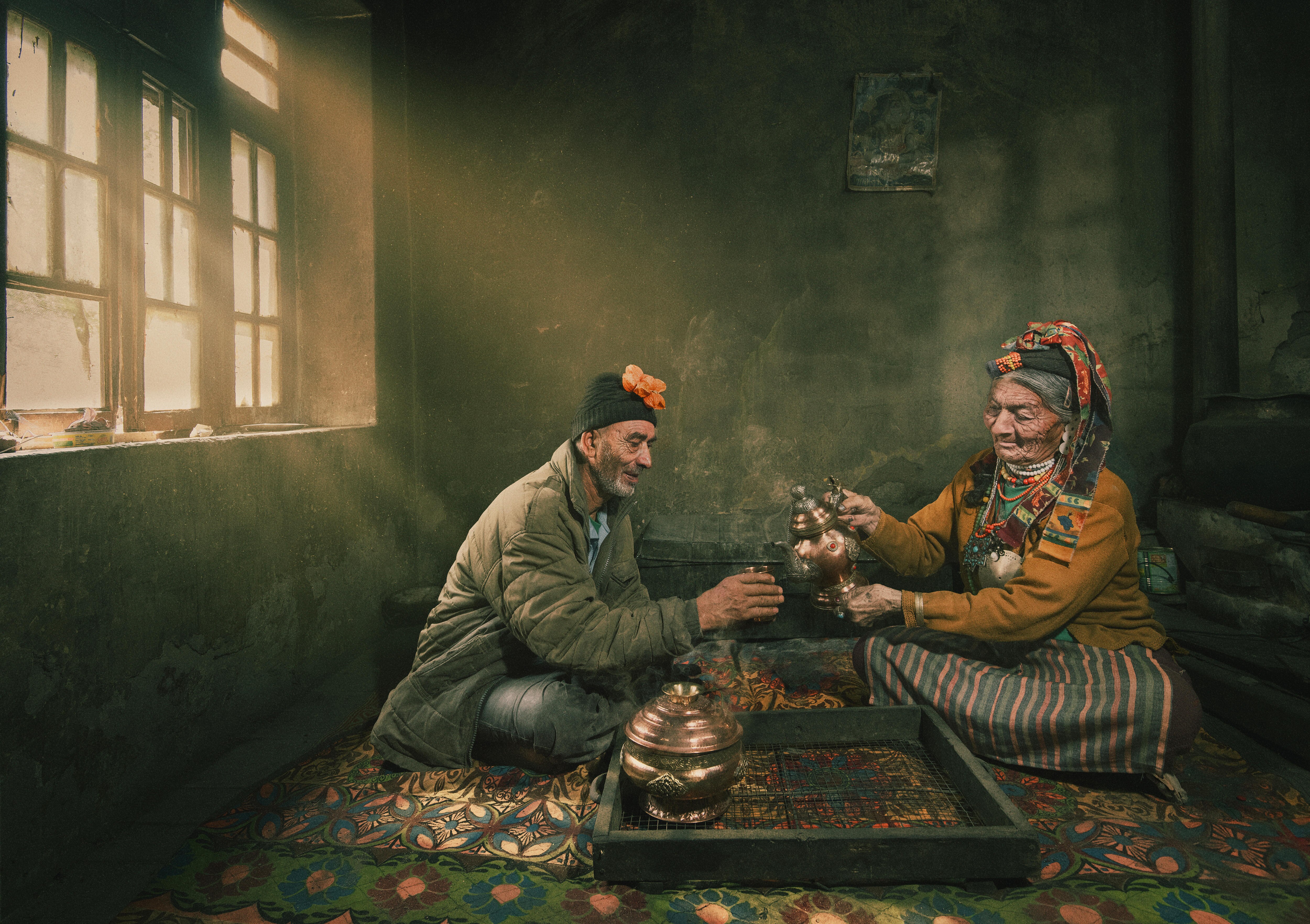 A man and woman sit cross-legged in a concrete room, sharing tea. They&#x27;re lit up by morning rays through a long window. 