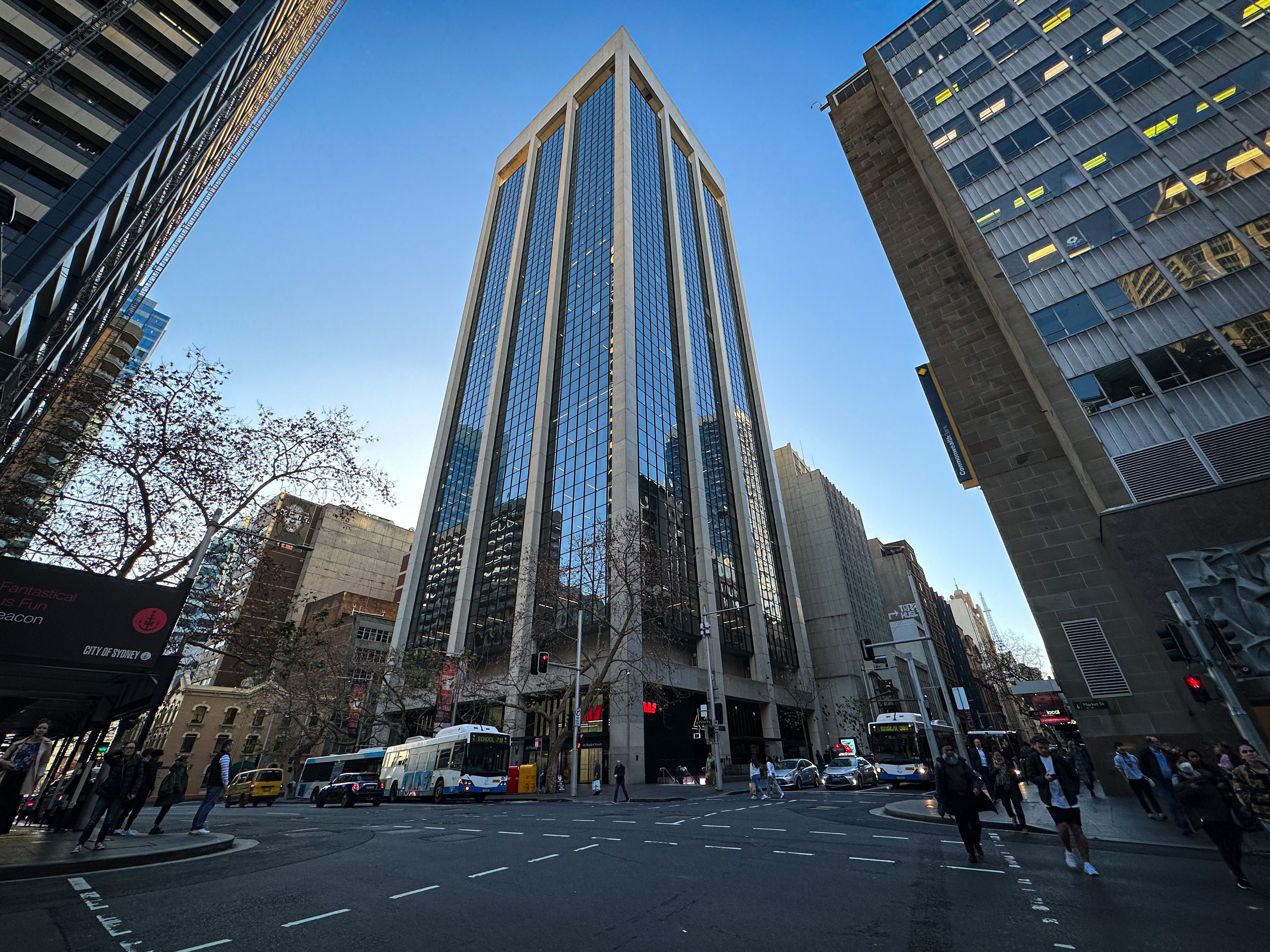 A tall office building taken in a central business district with buses and pedestrians nearby.