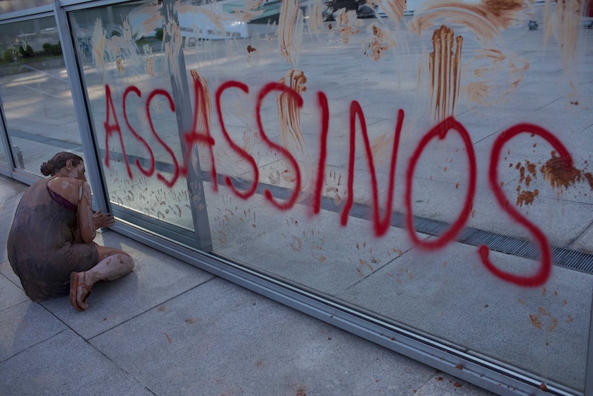 A woman covered in mud crouches next to a glass wall with "ASSASSINOS" painted in red. Muddy hand prints are all over the glass