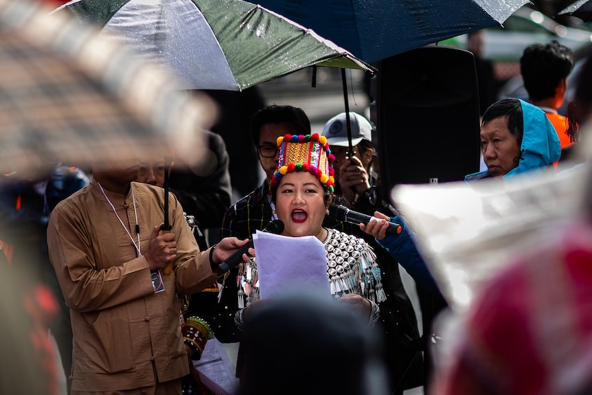A woman in a colourful traditional hat speaks into microphones surrounded by umbrellas. 