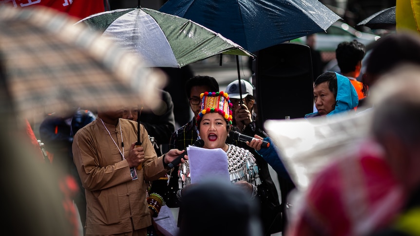 A woman in a colourful traditional hat speaks into microphones surrounded by umbrellas. 