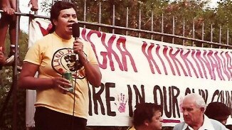 A woman with short dark hair standing on a garbage bin with a microphone in her hand in front of a protest banner.