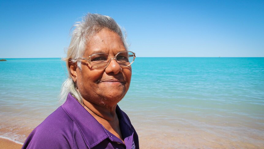 An Indigenous woman smiles at the beach looking out at crystal blue waters.