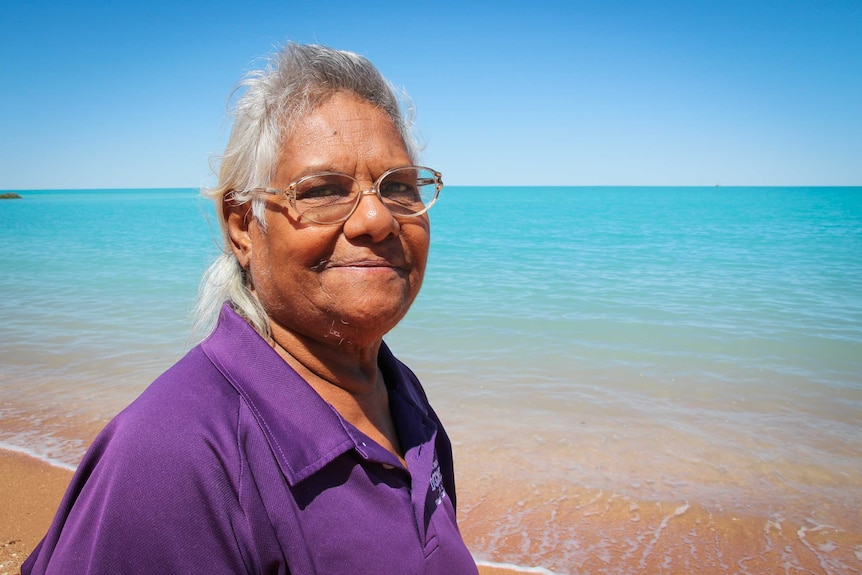 An Indigenous woman smiles at the beach looking out at crystal blue waters.