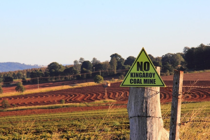A triangular 'no kingaroy coal mine' sign hooked to a fence, with a red soil background. 