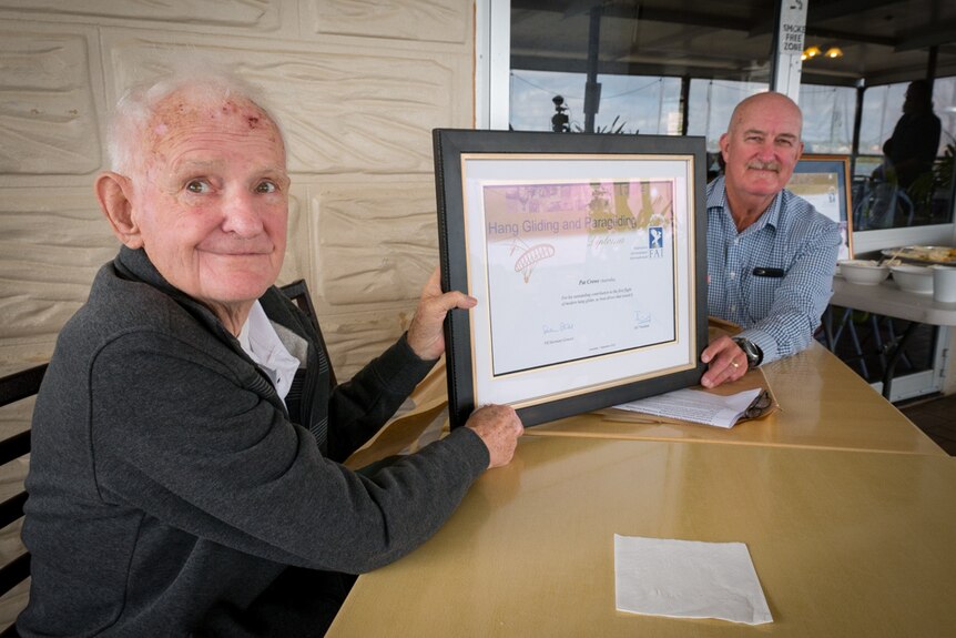 Elderly man holding a framed diploma with another man in the background.